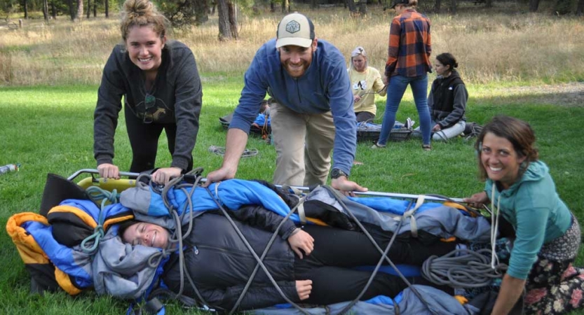 One person lies bound on a stretcher while three people roll them over and smile at the camera. They are all participating in a wilderness first responder course.